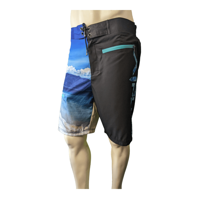 A mannequin displays board shorts from the Lava Flow line, featuring a split design: one half adorned with a scenic beach and ocean print, and the other in solid black with a turquoise zipper pocket. Perfect for beach apparel enthusiasts, these shorts are secured with a lace-up front.