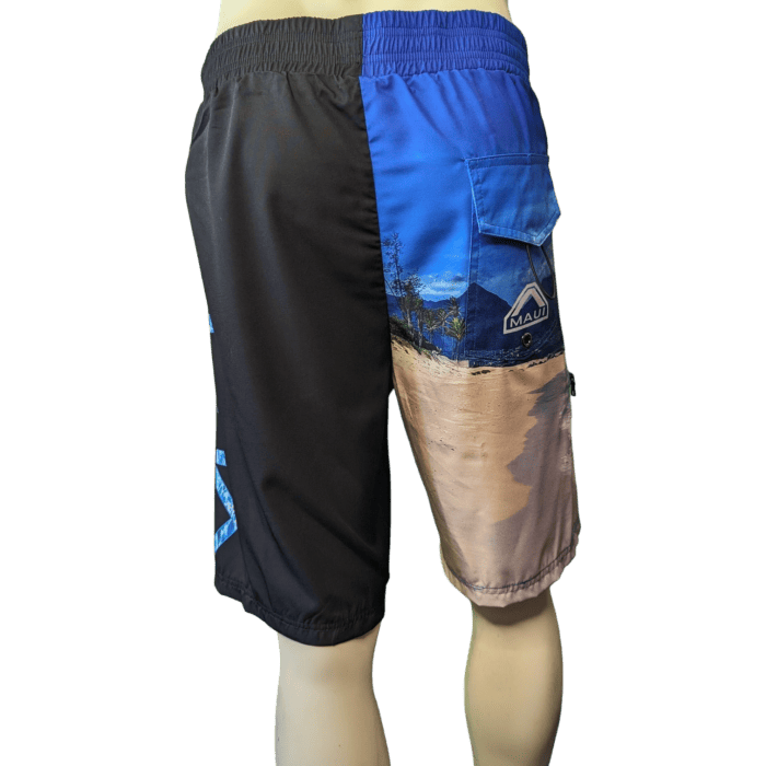 The Lava Flow beach apparel shorts are displayed on a mannequin, featuring a unique design with one half in solid black and the other half showcasing a Hawaii beach scene with trees, water, and a blue sky. The right side has a pocket adorned with an Ohana logo, while the left side has lettering near the hem.