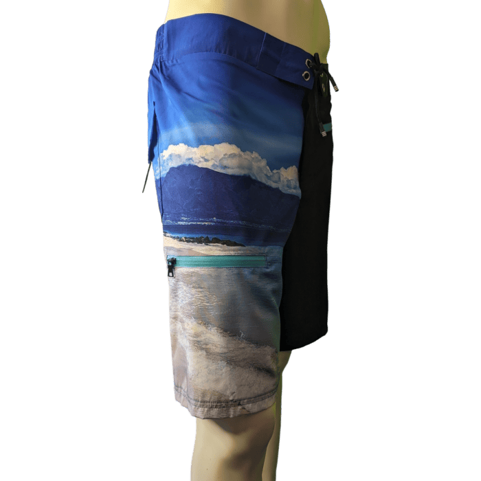 The mannequin is dressed in "Lava Flow" board shorts, showcasing a scenic landscape design of a Hawaiian beach, sea, and mountains beneath a blue sky with white clouds. These Aloha-inspired shorts come with a dark blue waistband and have a pocket on the right side. The image captures a side view of the shorts.