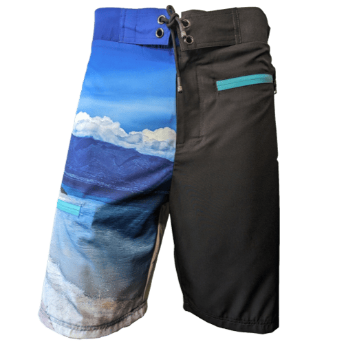 The Lava Flow board shorts feature a unique Alpha Maui design: the left leg showcases a scenic ocean and mountain landscape in shades of blue, while the right leg is solid black. These shorts are enhanced with turquoise zipper details on the pockets and have an adjustable drawstring waist, making them perfect Ohana beach apparel.