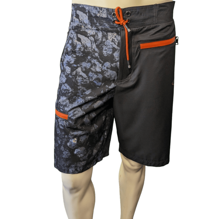 Image of a mannequin wearing men's board shorts. The left side features a gray camo design with an orange-trimmed pocket, while the right side is solid black with an orange-trimmed zipper pocket. Fastened with an orange drawstring, these Lava Flow shorts bring a touch of Aloha to your wardrobe.