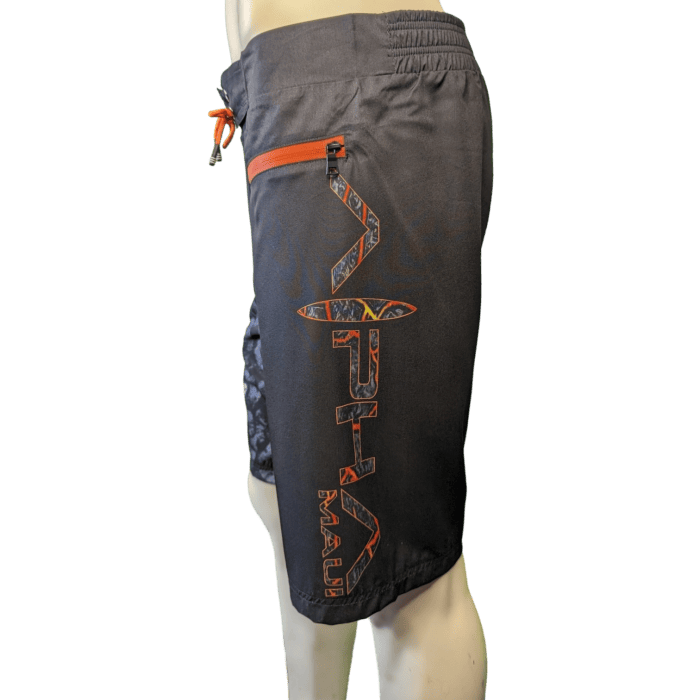 Side view of a person wearing the Lava Flow board shorts, showcasing a vibrant tribal pattern in shades of gray. These stylish shorts feature a red zipper pocket on the right side and orange writing that spells "Alpha Maui," adorned with an integrated surfboard graphic. The elastic waistband guarantees both comfort and style, making them perfect for any beach adventure.