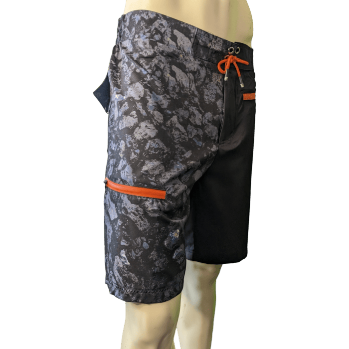 A mannequin showcases a pair of Lava Flow men's swim trunks in black and gray camouflage with striking orange accents. Inspired by Hawaii, these trunks feature orange zippers on the side pocket and an orange drawstring at the waist, bringing an Aloha vibe to your beachwear.