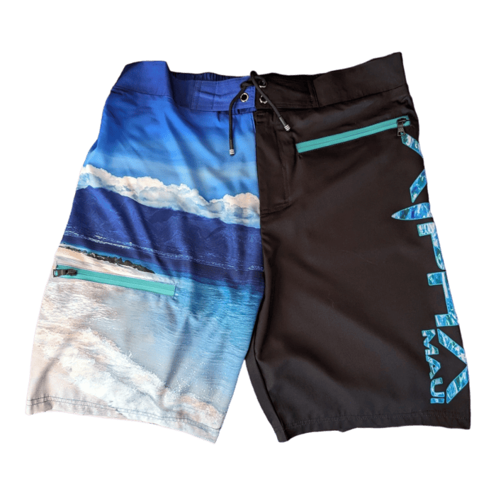 Introducing the Lava Flow board shorts. These shorts feature a split design; the left side boasts a vibrant beach scene with blue waters and a cloud-filled sky, while the right side is solid black adorned with a teal zipper and vertical teal text reading "Alpha Maui" along the leg. These are perfect for capturing that Aloha vibe in Hawaii.