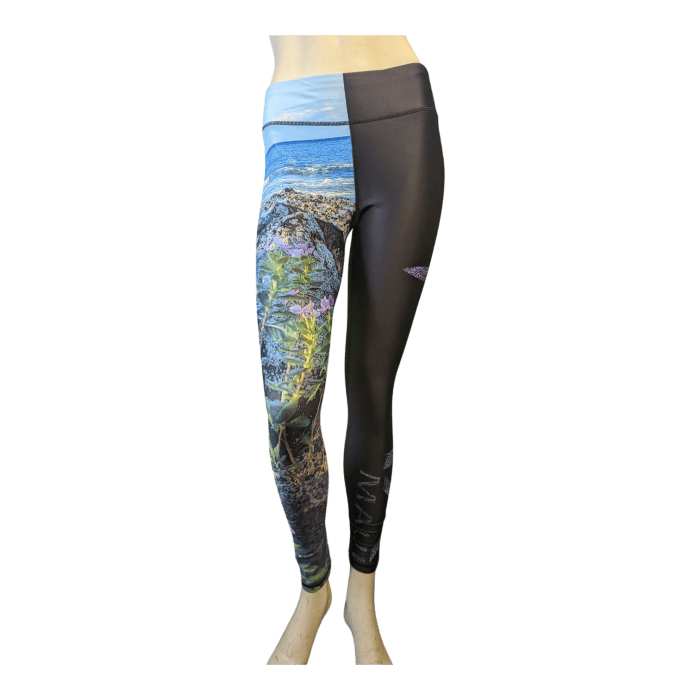 Introducing the Alpha Maui La Perouse leggings, a unique pair featuring dual-themed designs: the left leg displays a vibrant nature scene with rocks and flora by the sea, evoking the essence of Hawaii, while the right leg is solid black. The waistband combines light blue and black sections for added style. These distinctive leggings are showcased on a mannequin.