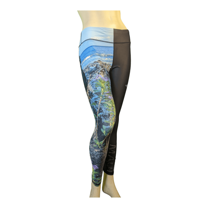 A mannequin is displaying Alpha Maui La Perouse leggings, available in 3/4 and full-length styles. These high-waisted leggings feature a unique Hawaiian-inspired design: the left leg boasts a detailed, vibrant nature scene while the right leg remains solid black. The waistband is adorned with an elegant light blue and gray pattern. The snug fit captures the essence and beauty of the Ohana spirit in Hawaiian culture.