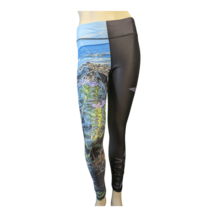 The mannequin is wearing Alpha Maui La Perouse leggings. The left leg showcases a vibrant nature scene with rocks, green plants, pink flowers, and a blue sky, while the right leg is solid black with an Alpha Maui logo on the thigh.