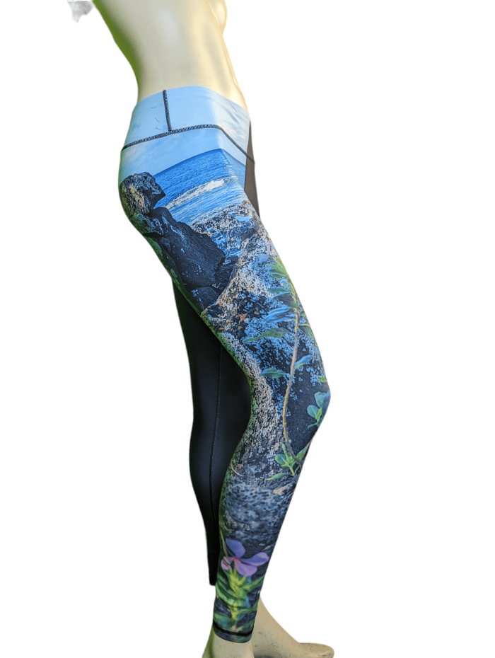 A side view of a person wearing colorful, form-fitting Alpha Maui La Perouse leggings featuring a nature scene with rocks, plants, and ocean waves. These leggings are perfect beach apparel. The rest of the leggings are black, and the image highlights the vibrant "Aloha" design on the left leg. The background is white.