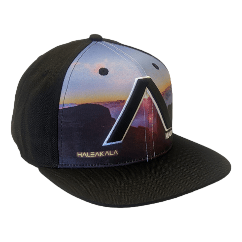 The Haleakala - Keiki Cap is a stylish black snapback designed specifically for kids. The cap features a vibrant graphic of a sunset landscape over mountains on the front panel, with "HALEAKALA" subtly written towards the bottom left. A prominent letter "A" adorns the crown. Ideal for beach outings, it beautifully captures the essence of Aloha and Ohana.