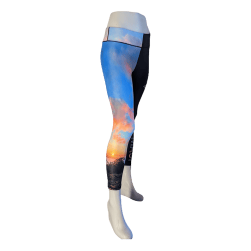 A mannequin wearing colorful leggings stands with its right leg forward. The leggings, part of our Alpha Maui Ulupalakua (3/4 Length) collection, display a vibrant scenic print featuring a sunset with pink, orange, and blue hues above dark mountains.