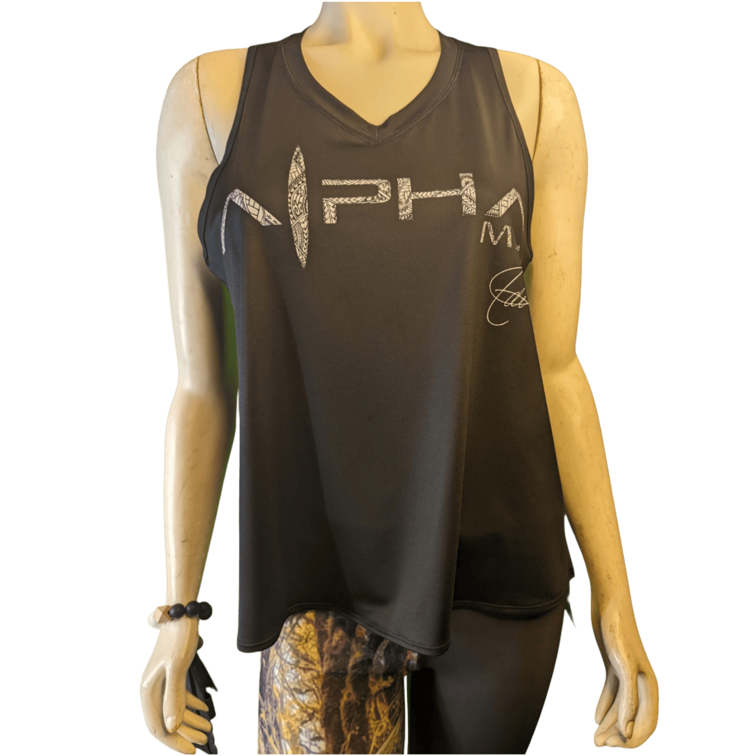 A mannequin is showcasing the Adi Tank Top, a black sleeveless athletic top featuring the word "Alpha" in a distressed white font across the chest. The mannequin exudes an Alpha Maui vibe and is accessorized with a bracelet and patterned leggings.