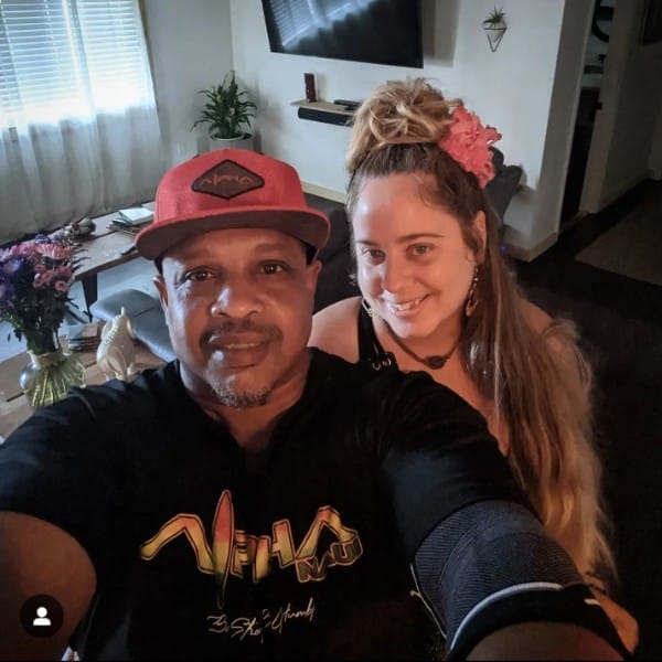 A man and woman smile for a selfie in a cozy living room with a large TV on the wall, plants, and a bouquet of flowers on a table. Embracing the spirit of Ohana, the man wears a black T-shirt and red cap, while the woman has long hair with a pink flower accessory and wears a black top.