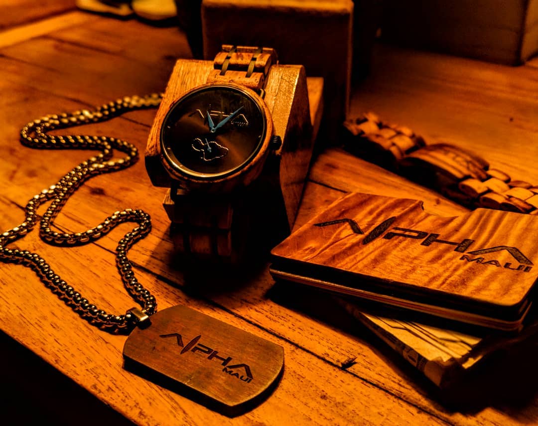 A wooden wristwatch is displayed on a wooden surface next to a dog tag on a chain, a wooden wallet, and a bracelet. All items feature the engraved brand name "APHA." The setting has a warm, golden-yellow glow, highlighting the craftsmanship and wood textures, evoking the relaxed vibe of Alpha Maui beach apparel.