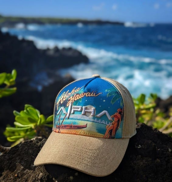 A colorful hat with the text "Aloha Hawaii" and "Alpha Maui" rests on a rocky surface with a scenic ocean backdrop. The hat features a vibrant design of palm trees, a sailboat, and a couple embracing. Green foliage and waves crashing against the rocks are visible in the background.