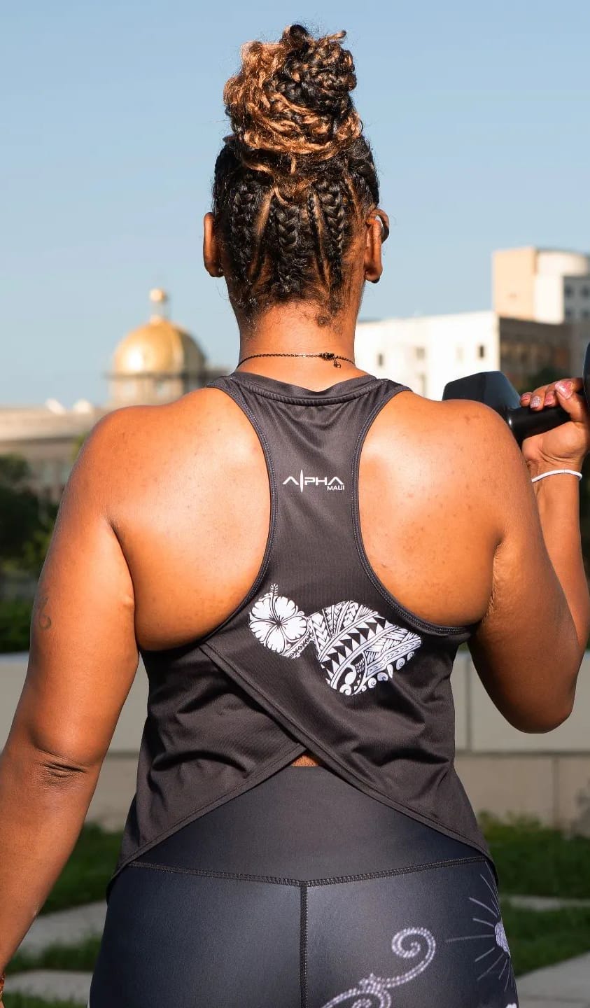 A person in workout attire, featuring a black tank top with a floral design from the Alpha Maui collection, stands with their back to the camera. They hold a dumbbell in their raised right hand. The background includes a cityscape with a domed building.