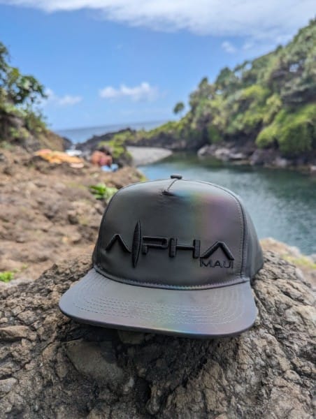 A gray baseball cap with "Alpha Maui" embroidered on it rests on a rock. In the background, there is a scenic view of a rocky coastline and lush greenery under a partly cloudy sky, embodying the spirit of Hawaii. This beach apparel piece exudes that perfect Ohana vibe.