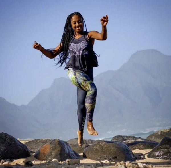 A person with long braided hair is joyfully jumping over rocks at the beach in Hawaii. They are wearing a black tank top and colorful leggings. Mountains and a clear blue sky form the background, creating a scenic outdoor setting reminiscent of an Alpha Maui adventure with friends and family, or 'Ohana'.