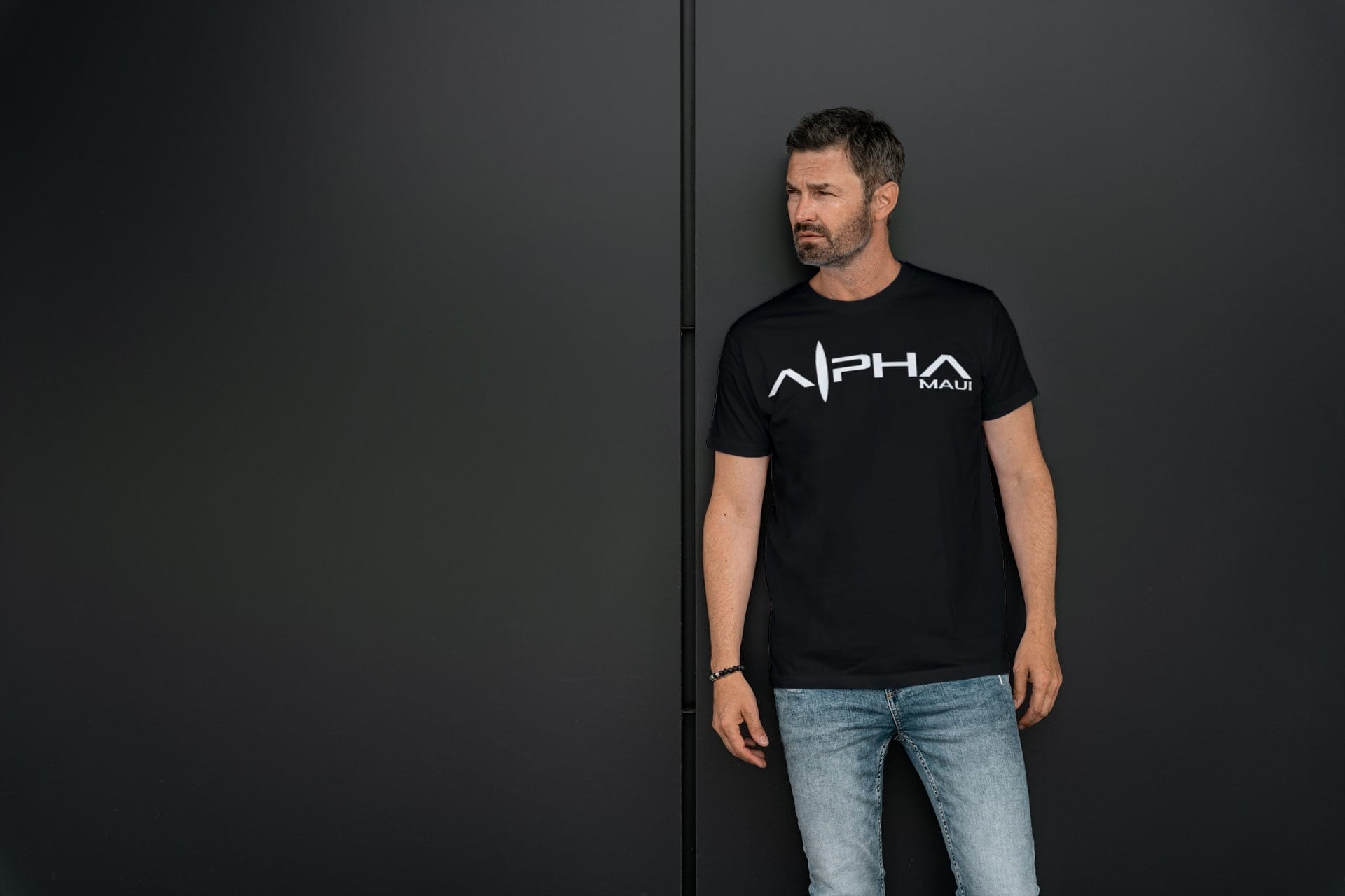 A man with short dark hair and a beard stands against a dark wall. He is wearing a black T-shirt with the text "Alpha Maui" in white letters and light blue jeans, embodying beach apparel. He looks to the side with a serious expression, evoking a sense of Hawaii's laid-back yet focused vibe.