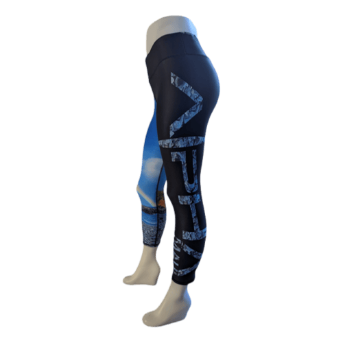 A mannequin displays the Alpha Maui Haukini Bay (3/4 Length) leggings, featuring black fabric with striking blue accent patterns and "LEVEL" printed in large white letters along the side. Ideal for beachwear, these leggings boast a scenic blue and white design on the lower part of the left leg, embodying that Ohana spirit.