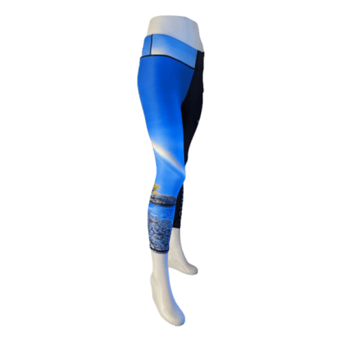 The image features a mannequin showcasing Alpha Maui's Haukini Bay beach apparel: 3/4 length leggings with a stunning landscape design. The leggings transition from dark to light blue, depicting the sea, sky, and a small island or coastline, embodying the serene essence of Ohana.