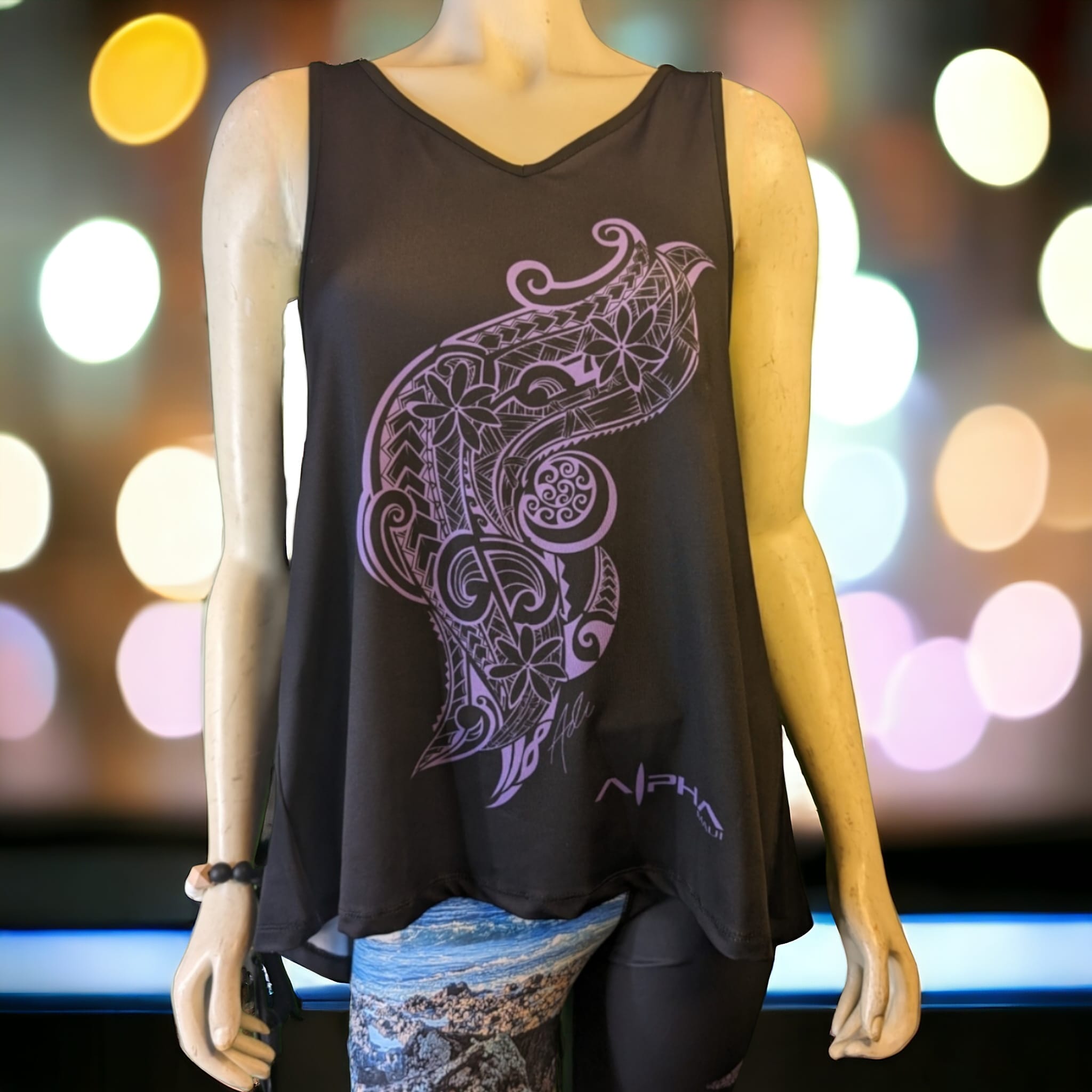 A mannequin is dressed in a dark sleeveless top featuring a large, intricate design in pink on the front, reminiscent of beach apparel. The design resembles a stylized feather or tribal pattern, perfect for an Alpha Maui look. The mannequin also wears blue patterned leggings. The background is blurred with colorful bokeh lights.