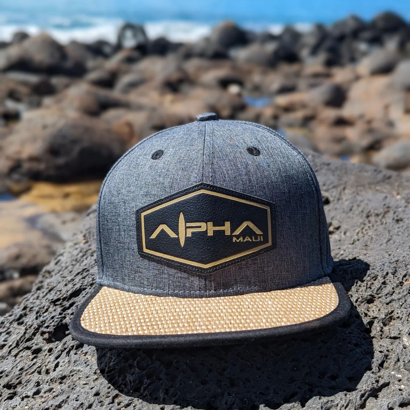 A gray and tan baseball cap with a hexagonal patch that reads "Alpha Maui" rests on a large rock, set against the blurred backdrop of a rocky beach and the serene ocean in Hawaii. Perfect for your beach apparel collection.