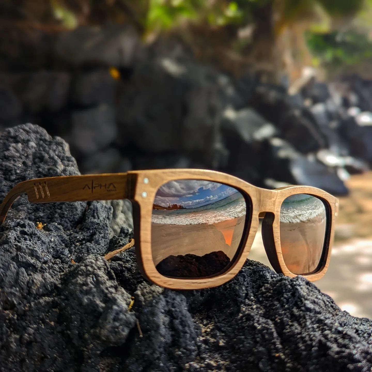 A pair of Alpha Maui wooden sunglasses resting on a textured black rock. The lenses reflect a serene beach scene with waves gently lapping the shore and distant mountains under a clear blue sky. The surroundings are slightly blurred, emphasizing the beach apparel sunglasses.