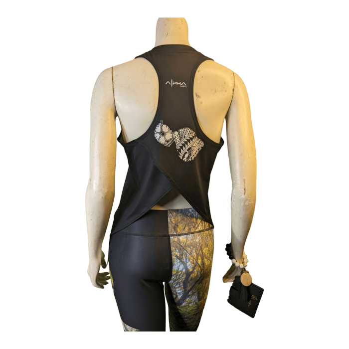 The image depicts a mannequin from the back wearing the "Alpha Maui U'i Tank," a black sleeveless top with an open-back design, featuring a butterfly pattern. The mannequin is paired with fitted leggings showcasing a forest print and is holding a small black pouch in the left hand, exuding an Aloha vibe.