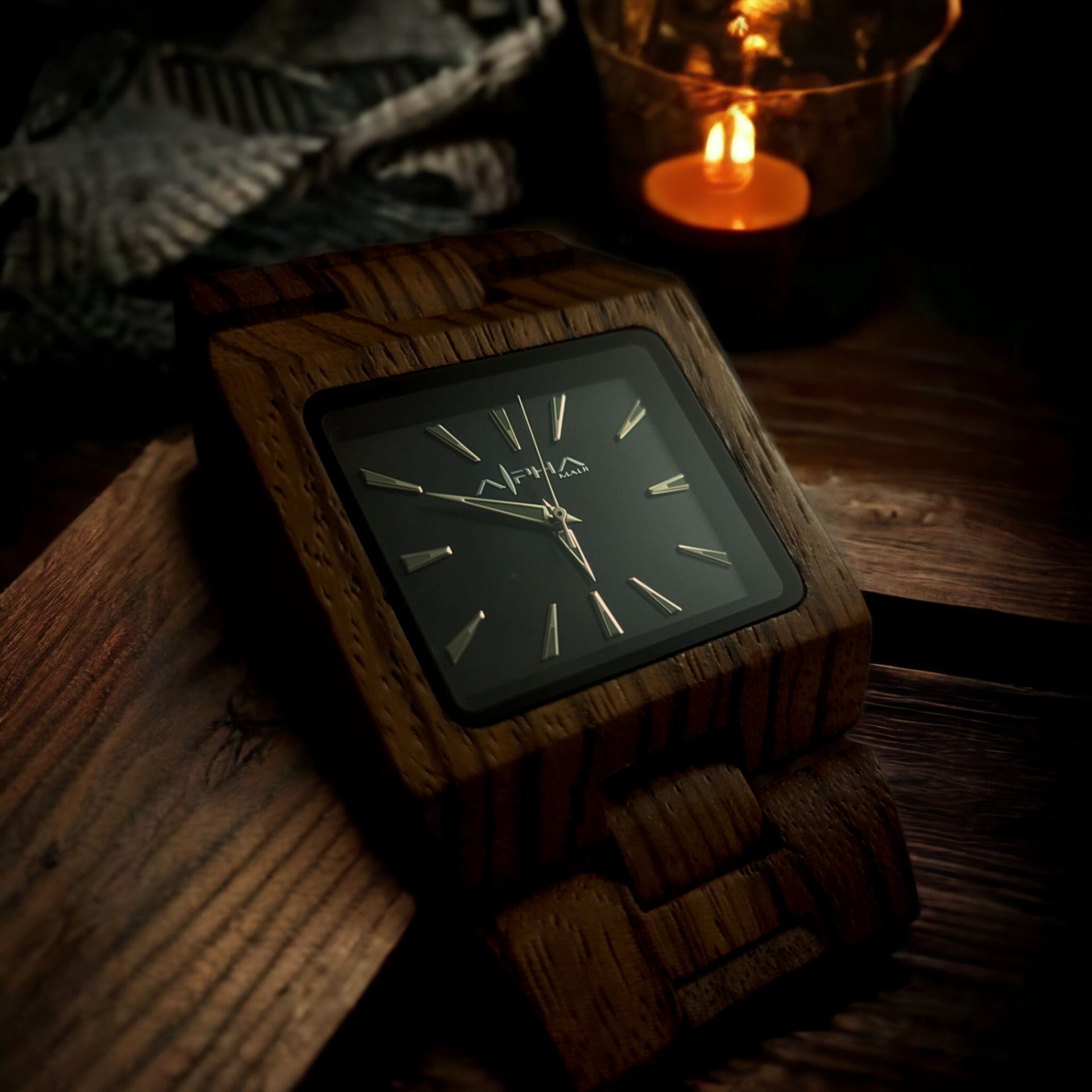 A wooden wristwatch with a black square face and gold hour markers rests on a wooden surface. A lit candle in a glass holder can be seen in the background, casting a warm, soft light and creating an Aloha atmosphere filled with cozy Ohana vibes.