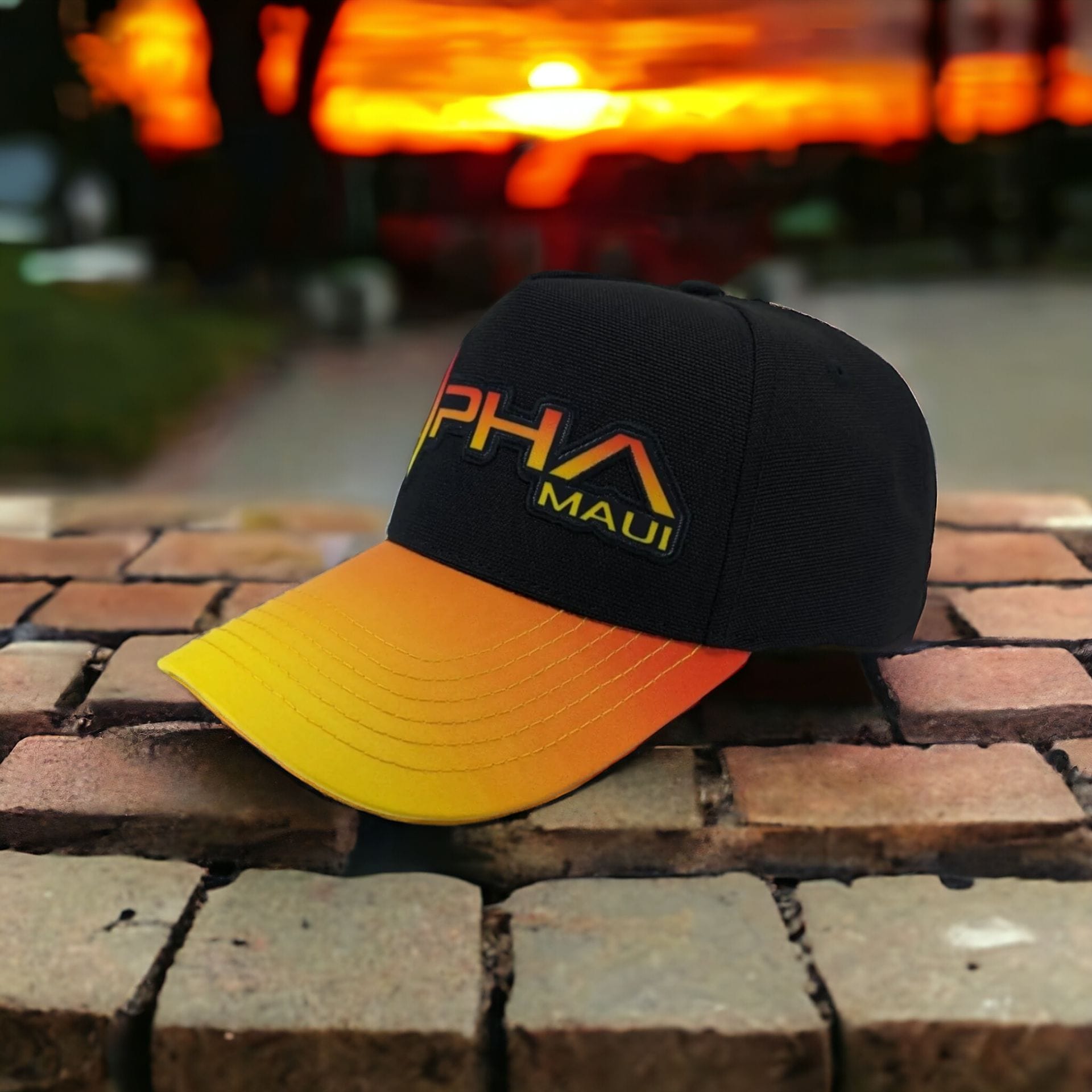 A black and yellow gradient baseball cap with "Alpha Maui" embroidered on it is placed on a brick surface. The background features a vibrant, colorful sunset with deep orange and yellow hues and blurred, shadowy trees, evoking the spirit of Hawaii's Ohana.