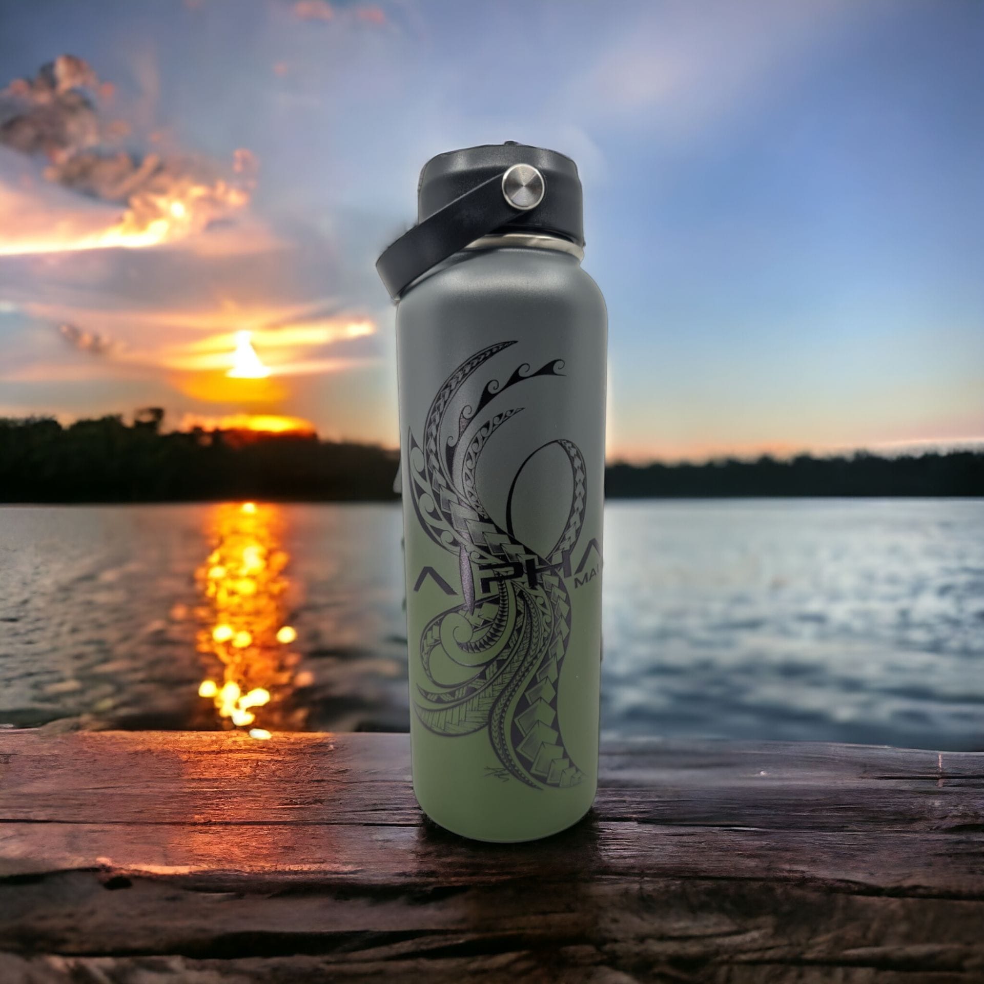 A reusable water bottle with intricate black designs sits on a wooden surface overlooking a lake. The sun sets in the background, casting an orange and purple hue across the sky and reflecting on the water, creating an Aloha-inspired scene.