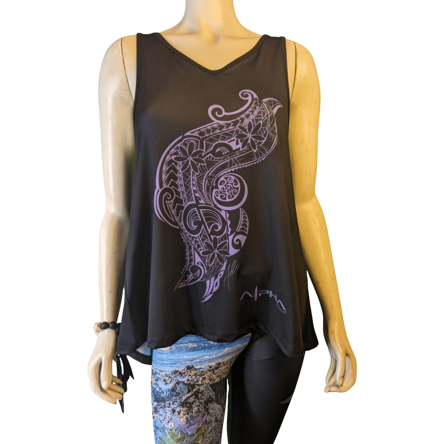 A mannequin wearing the Alpha Maui U'i Tank, a sleeveless V-neck top adorned with intricate black and purple patterns reminiscent of Hawaii's vibrant culture. The tank is paired with colorful, landscape-patterned leggings. The mannequin has an "Aloha" bracelet on its left wrist, completing the Ohana-inspired look.