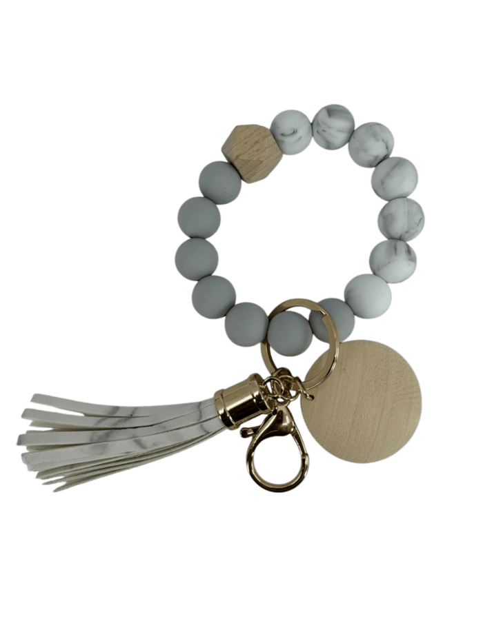 The Alpha Wristlet is a circular bracelet keychain with white marbled and wooden beads, a gold clip, a beige wooden medallion, and a white tassel featuring marbled accents.
