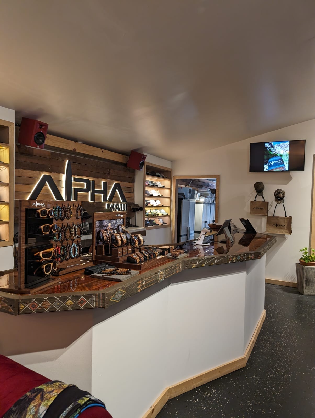 A trendy retail shop interior with wooden counter displays featuring various sunglasses and accessories. The backlit store sign reads "Alpha Maui." The walls have shelves with additional items, including beach apparel, and there's a small TV and wall decor evoking Hawaii on the right side.