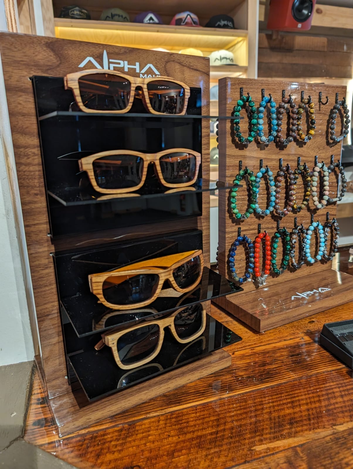 A wooden display stand features three pairs of Alpha Maui wooden-framed sunglasses on the left and an assortment of beaded bracelets hanging on hooks on the right. The setup is arranged neatly on a wooden countertop with a modern, rustic Aloha aesthetic.