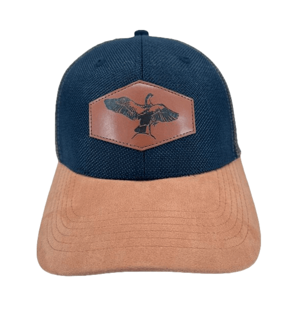 The Alpha Maui NeNe, a dark blue baseball cap with a light brown suede brim, is perfect for beach apparel. It features a brown leather patch on the front with a black silhouette of a goose in flight. The simple and outdoorsy design embodies the Ohana spirit.