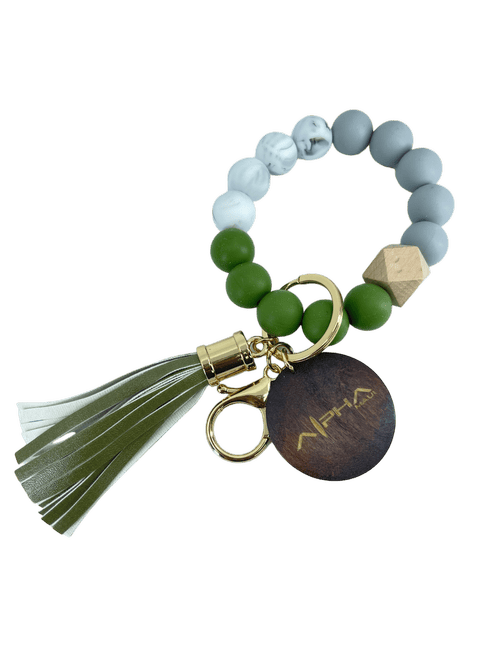 The Alpha Wristlet is a stylish keychain featuring a loop of round beads in white, green, and gray with a wooden geometric bead. It comes with a gold keyring and includes a dark wooden tag engraved with "Alpha Maui," along with a green and white tassel.