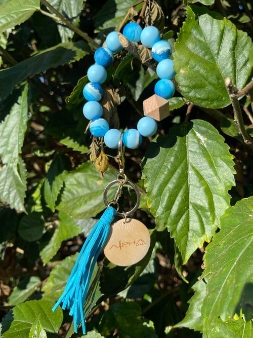 An Alpha Wristlet hanging from a green leafy plant. The wristlet showcases round blue beads, a wooden bead, a small wooden disc etched with an "Ohana" logo, and a blue tassel fastened to a metal ring. The background is lush with dense green foliage.