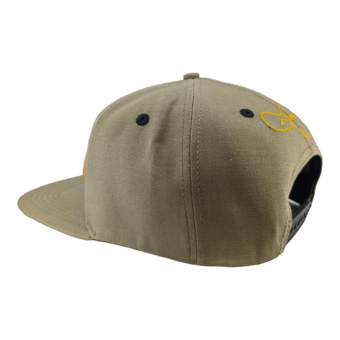 Side view of a beige baseball cap from Alpha Maui's Coalition Forces collection, featuring a yellow embroidered logo on the side. The cap boasts black vent holes and a black adjustable snapback closure, displayed against a plain black background.