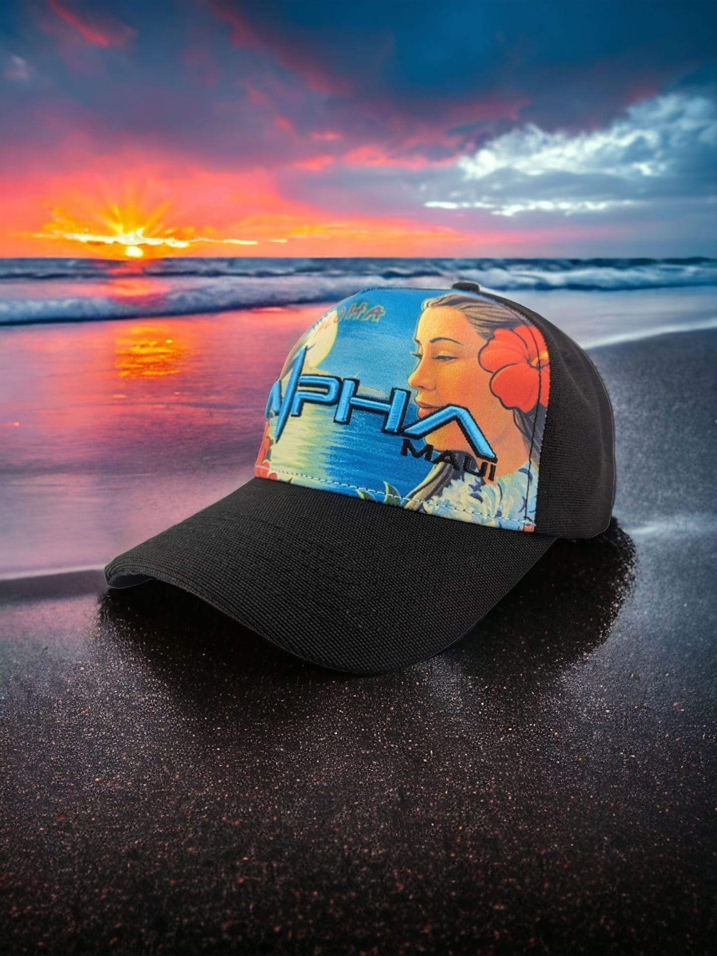 A black cap with a colorful design of a woman with a red flower in her hair and the word "Alpha" printed in bold letters on the front panel lies on a dark sandy beach at sunset. This piece of Hawaii-inspired beach apparel perfectly complements the serene backdrop created by the vibrant sky and calm sea.