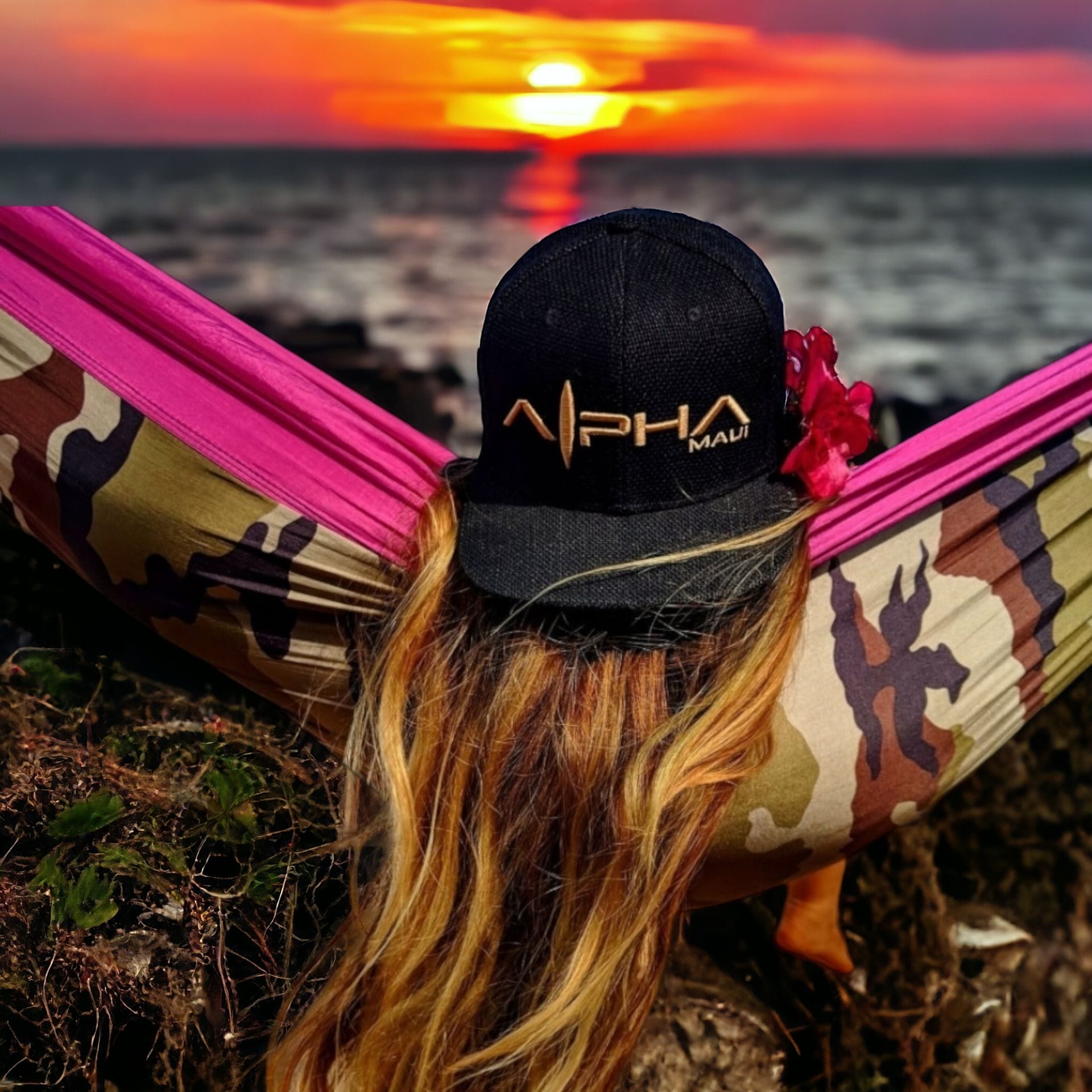 Person with long blonde hair wearing a black hat with "Alpha Maui" lettering, lying in a colorful pink and camo hammock. They are watching a vibrant sunset over a calm ocean, captured perfectly by their beach apparel. The scene is tranquil and scenic, an embodiment of true Aloha spirit.