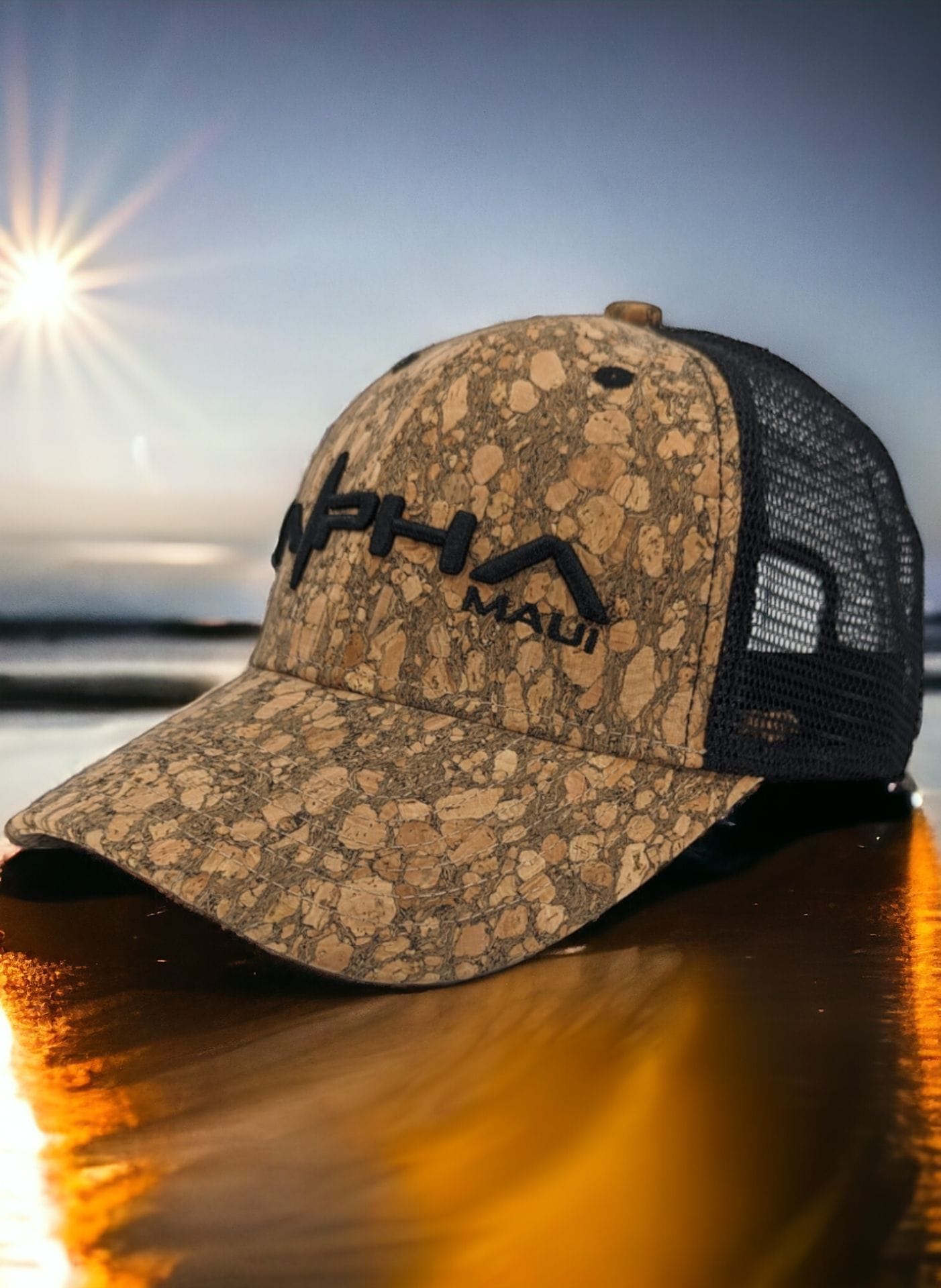A camouflage baseball cap with mesh backing featuring the word "Alpha" in bold black letters is placed on a reflective surface. The background showcases a serene Hawaiian landscape with a bright sun low in the sky and subtle water reflections, perfect for beach apparel enthusiasts.