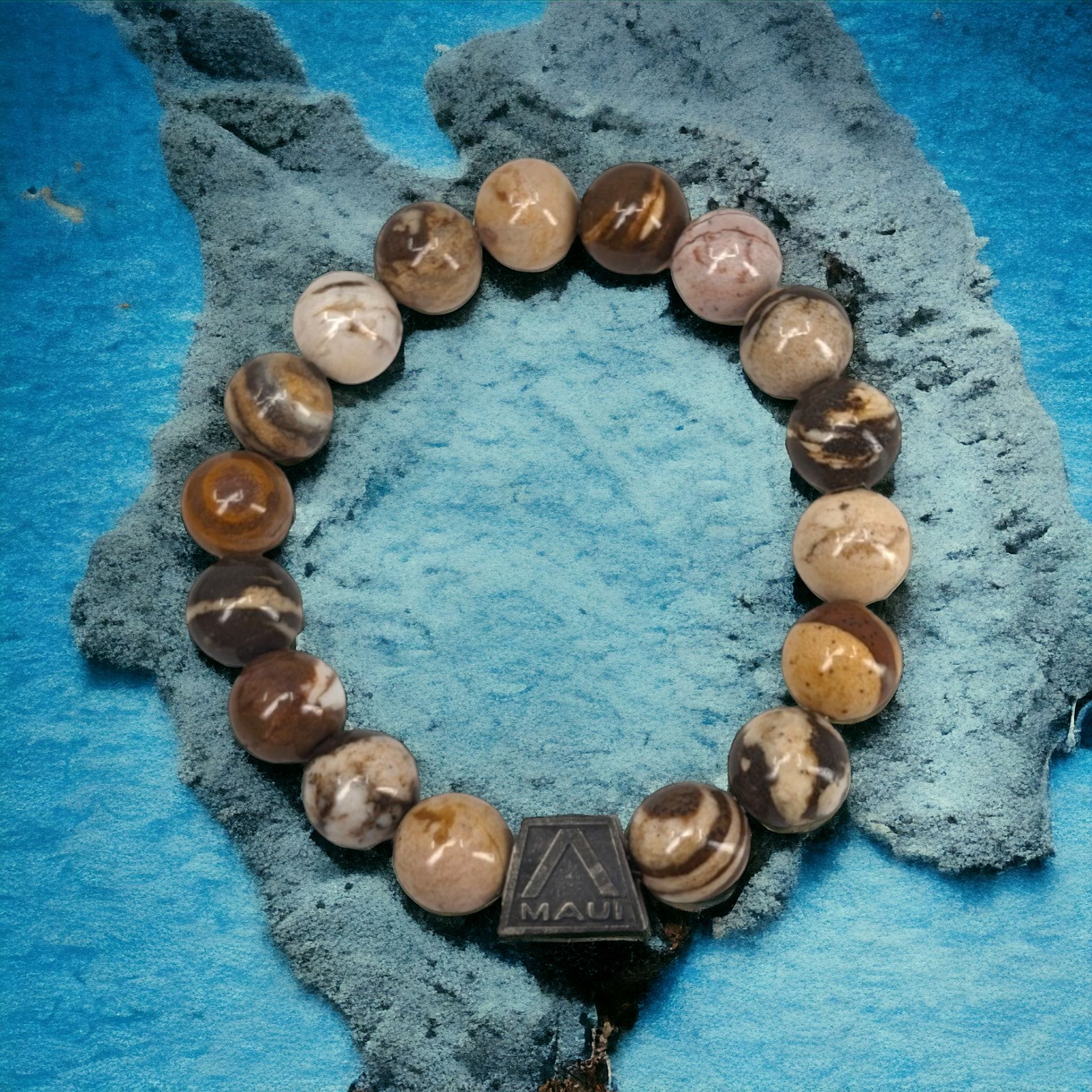 A beaded bracelet with round, marbled stones in various shades of brown and beige is displayed on a textured blue background. The bracelet features a small, dark brown rectangular charm inscribed with "Maui" and an 'A' logo, epitomizing Alpha Maui's beach apparel spirit.