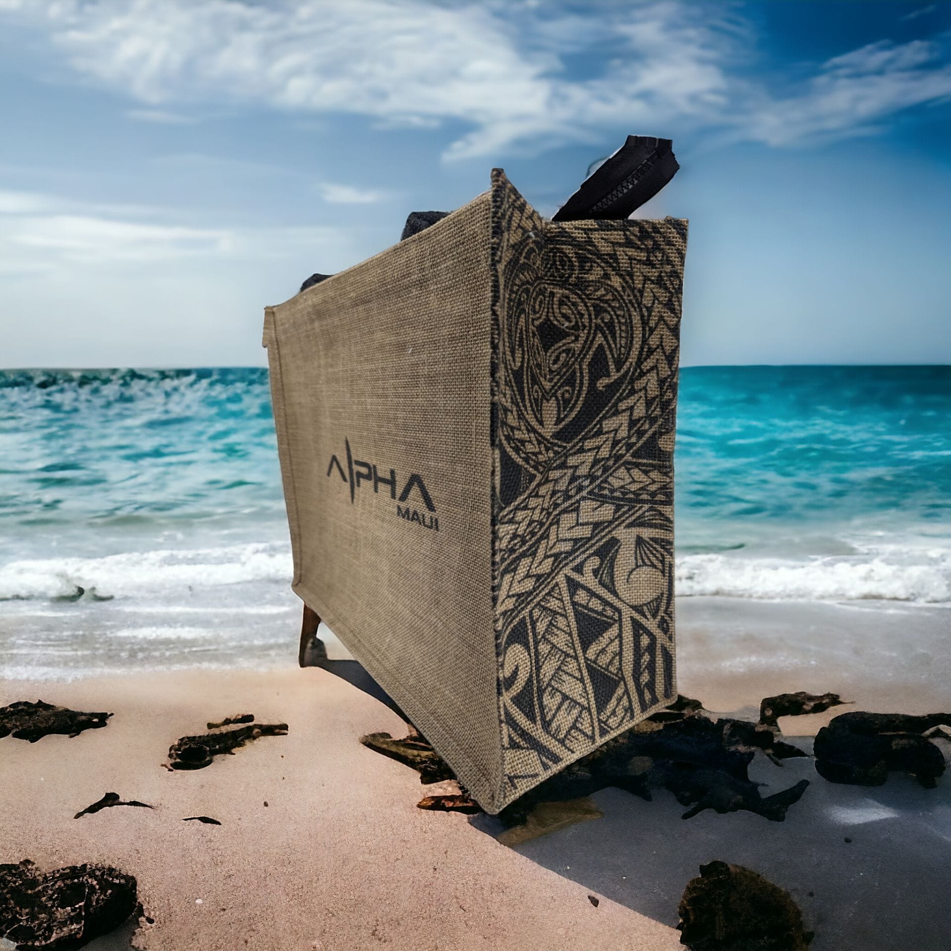 A large, intricately designed burlap bag with dark patterns and the text "Alpha Maui" stands upright on a sandy beach. The ocean waves are gently crashing in the background, and the sky is bright with scattered clouds, adding an extra touch of Aloha spirit to the scene.