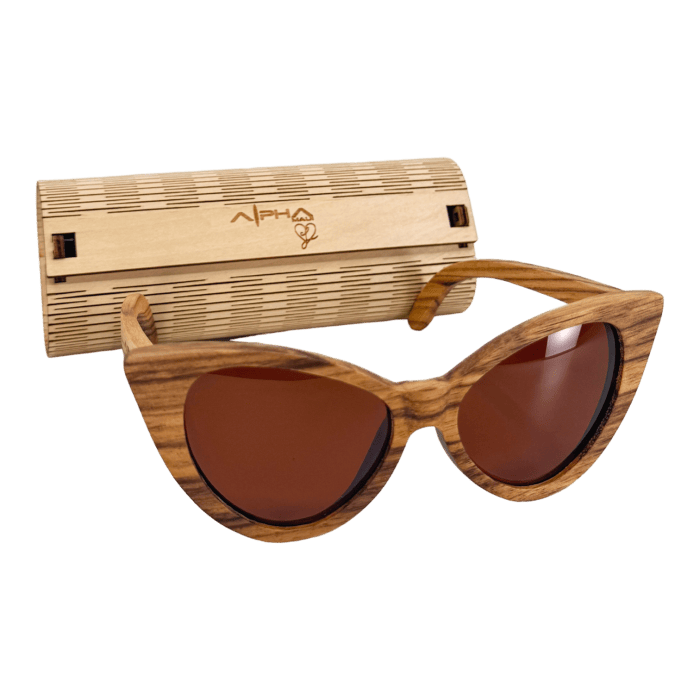 The Alpha Cat Eye sunglasses, featuring stylish wooden cat-eye frames and reddish-brown tinted lenses, are displayed in front of a bamboo case engraved with the Alpha logo. Both the sunglasses and case, embodying the spirit of Hawaii, are set against a black background.