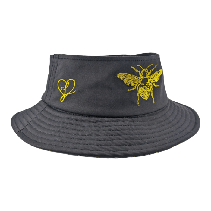 The Alpha Maui Bee Bucks is a black bucket hat with a neon yellow embroidered design, ideal for beachwear. One side showcases a bee detailed with intricate patterns, while the opposite side displays a simple heart shape with a small loop at its bottom. A quilted pattern along the brim brings an added touch of Aloha spirit.