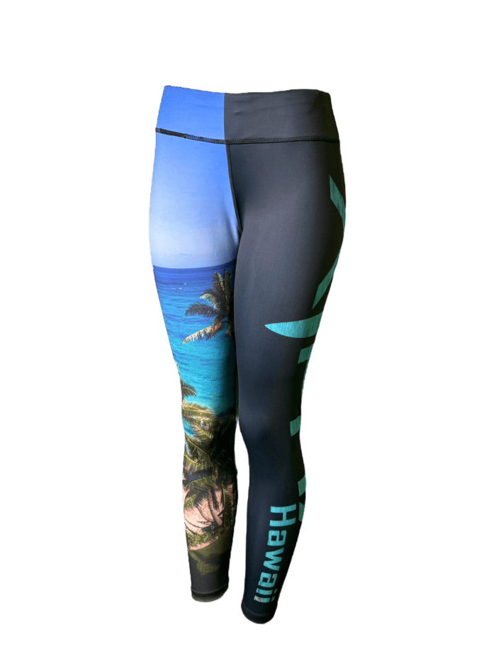 Introducing the Alpha Maui Waimanalo full-length leggings, featuring a distinctive split design. One leg beautifully showcases a tropical beach scene with palm trees and clear blue water, making it an ideal choice for beachwear. The other leg is solid black with aqua-colored text that reads "Hawaii," set against a black background.