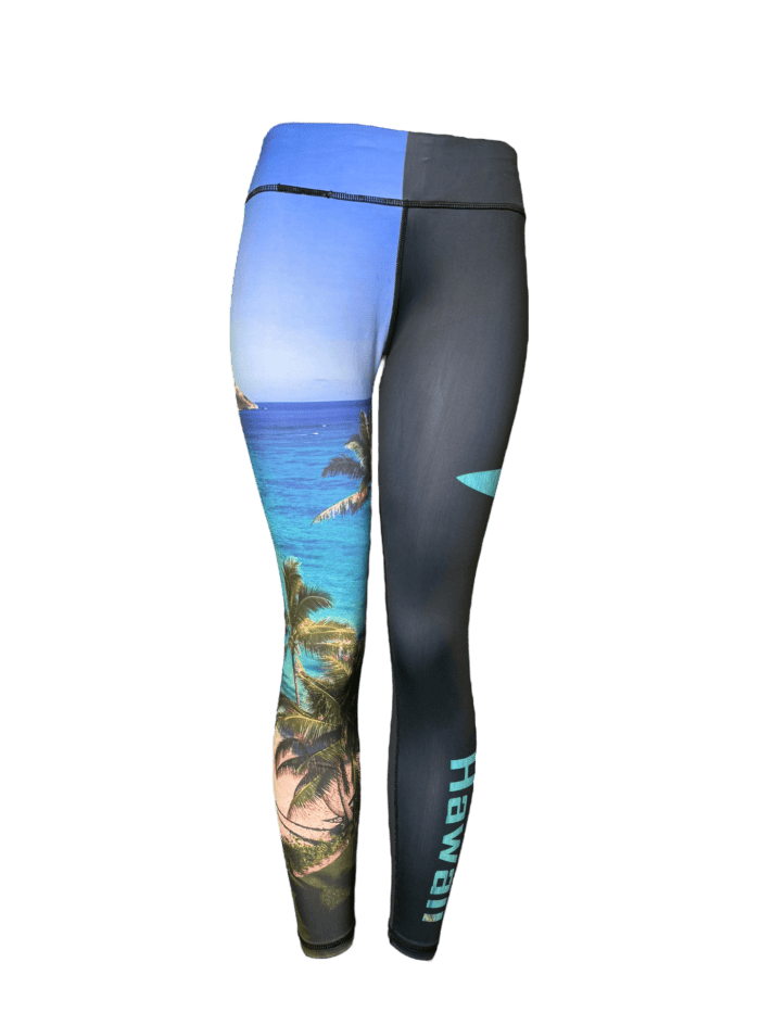 Introducing the Alpha Maui Waimanalo (Full Length) leggings, featuring a vibrant tropical beach scene on one leg and a plain dark design on the other. The word "Hawaii" is printed vertically on the dark leg, making these ideal for your beach apparel collection. The beach scene showcases a clear blue sky, palm trees, and turquoise water.