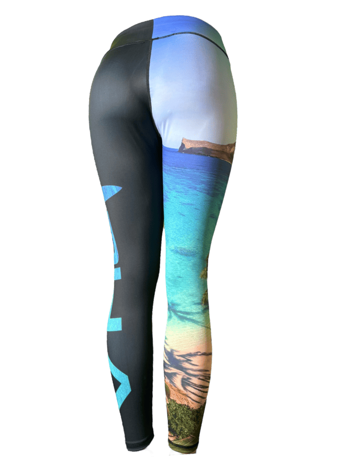 A person modeling the Alpha Maui Waimanalo full-length leggings: the left leg is solid black with white text, while the right leg features a vibrant Hawaii beach scene with blue water, sandy shores, and green foliage. The fabric stretches to fit snugly, showcasing the pattern perfectly.