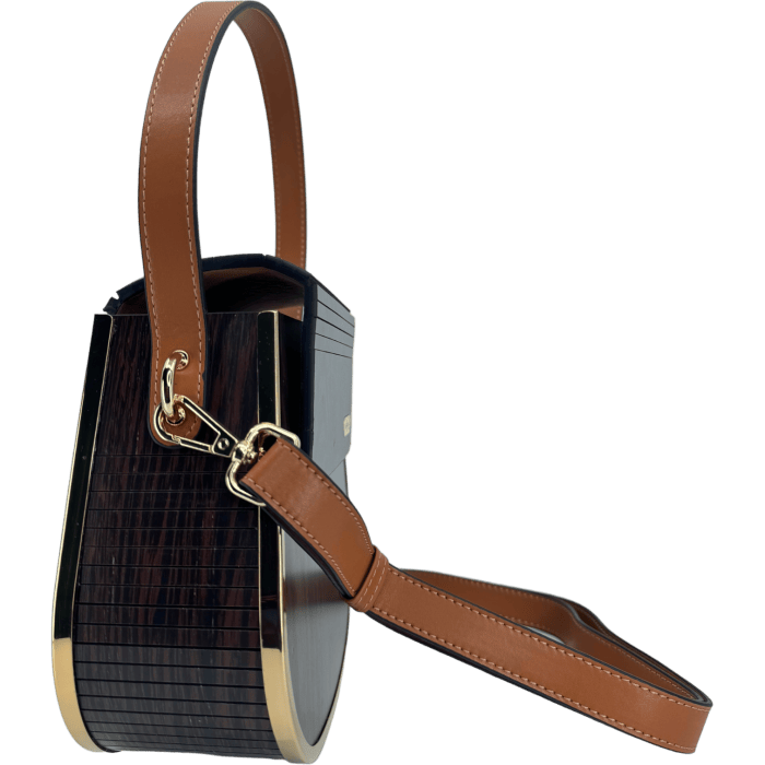 The Alpha Maui Rockit Carrier Purse is a stylish handbag perfect for your beach apparel, featuring a brown leather strap and gold hardware. This piece boasts a sleek, structured design with dark, wood-like textured sides and smooth brown leather accents. Its modern and elegant look is completed by a detachable strap, making it an ideal choice for an Ohana getaway in Hawaii.